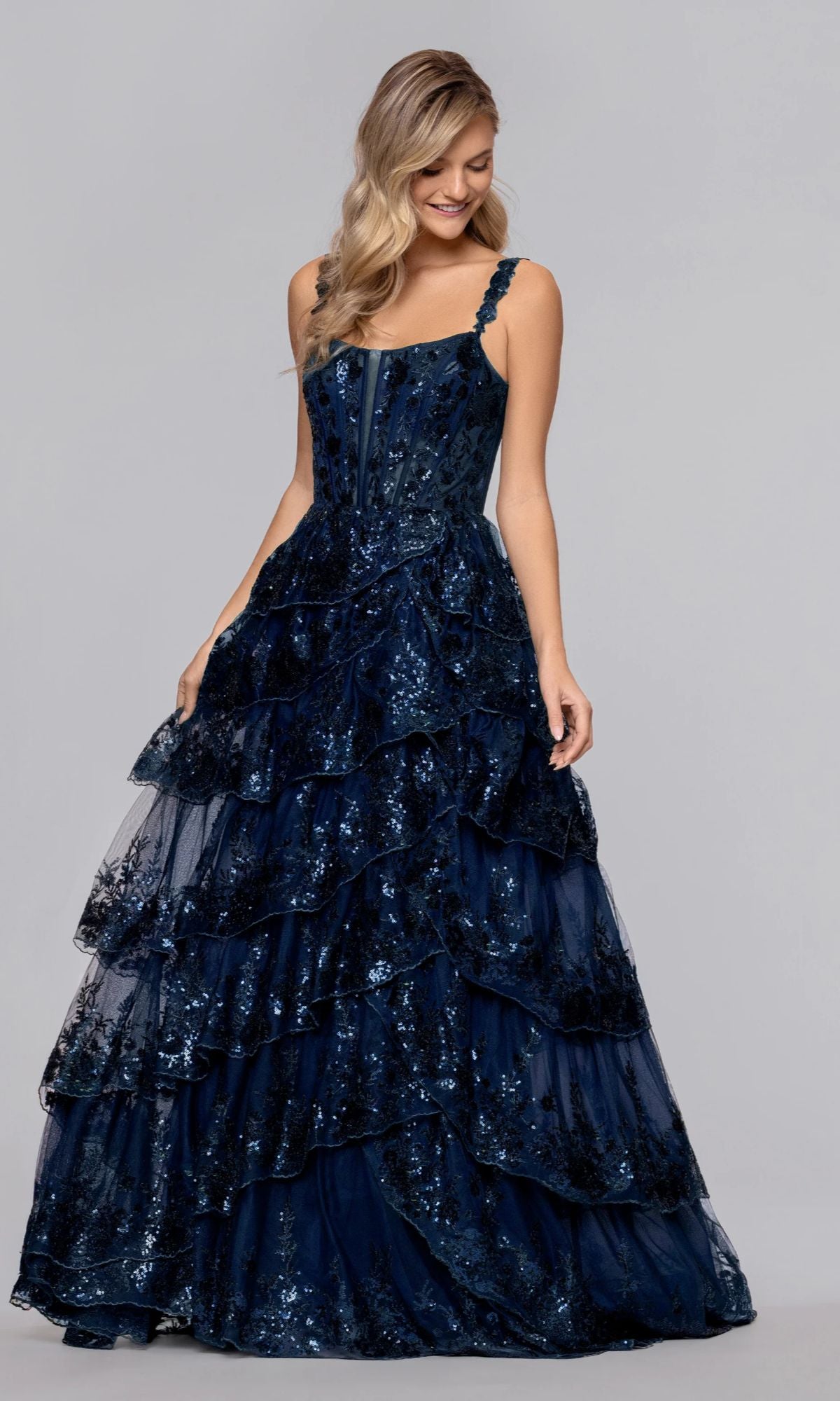 A woman in the Blondie Nites 6126BN long navy prom dress with sequin embellishments and a tiered A-line skirt stands against a plain gray background.